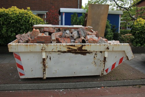 A large, white industrial skip bin filled with a mixture of red bricks, concrete debris, and broken construction materials, situated on a paved street in an urban residential area. The skip shows signs of rust and dirt, particularly around the top edge and sides, indicating frequent use. It is positioned directly on the pavement, with a red and white striped reflective warning strip running along its lower edge. In the background, there are green shrubs and trees, a blue shed, and residential buildings with red brick facades, suggesting a typical local neighbourhood. The scene appears to be daytime with natural light illuminating the area, consistent with waste removal activities commonly handled by companies such as Commercial Waste Tower Hamlets, serving the Tower Hamlets region. The debris in the skip is partly spilling over the top, reflecting a recent clearance or renovation project, typical of rubbish removal services in London and the surrounding area.
