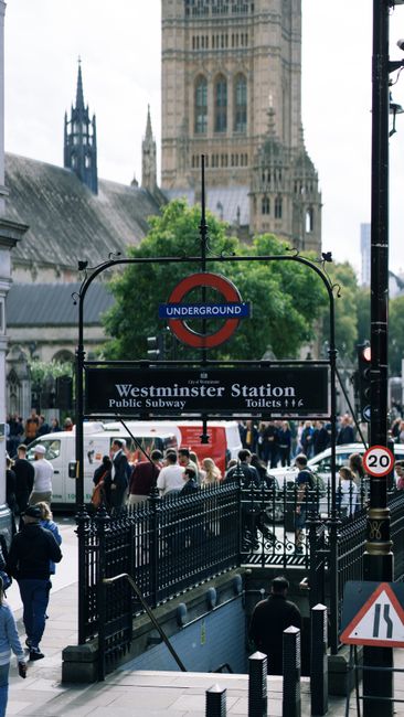 A busy scene outside Westminster Underground Station in London, featuring a black metal staircase leading down to the subway entrance, which is topped by a classic Underground sign with a red circle a